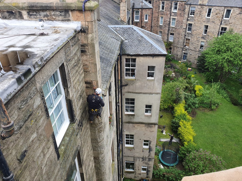 A worker in safety gear is abseiling down the side of a building, with a garden and trampoline visible below.