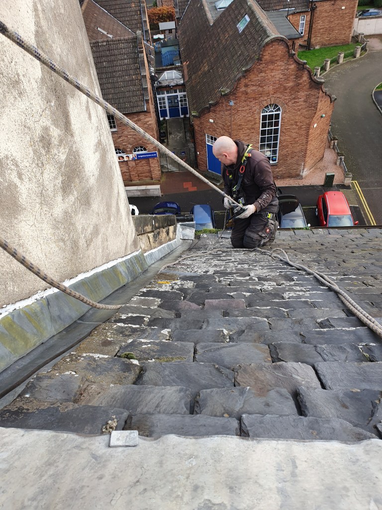 A person in safety gear repelling down a sloped roof, with a view of buildings and parked cars below.