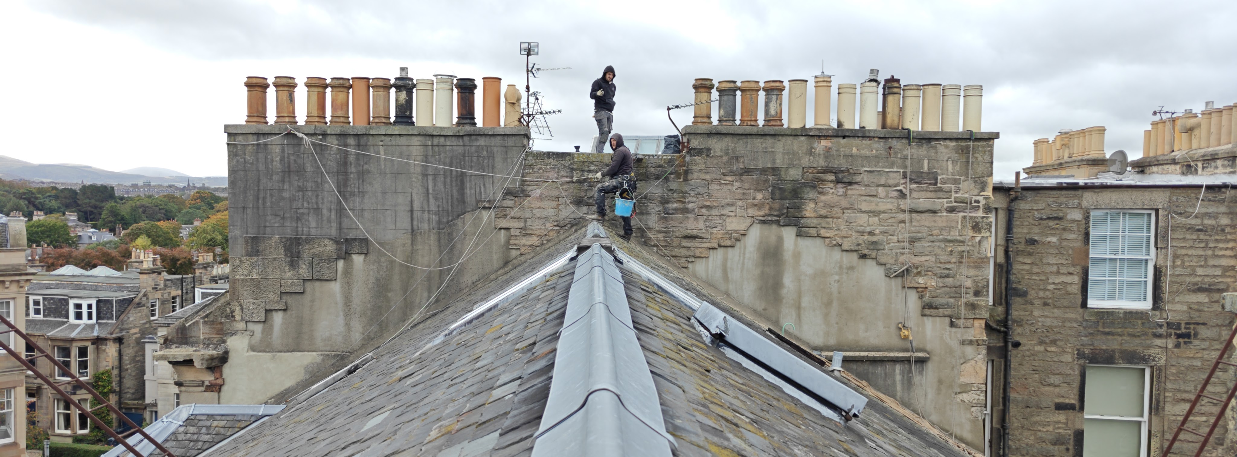 Two workers on a roof edge, with a view of rooftops and chimneys in the background, under a cloudy sky.