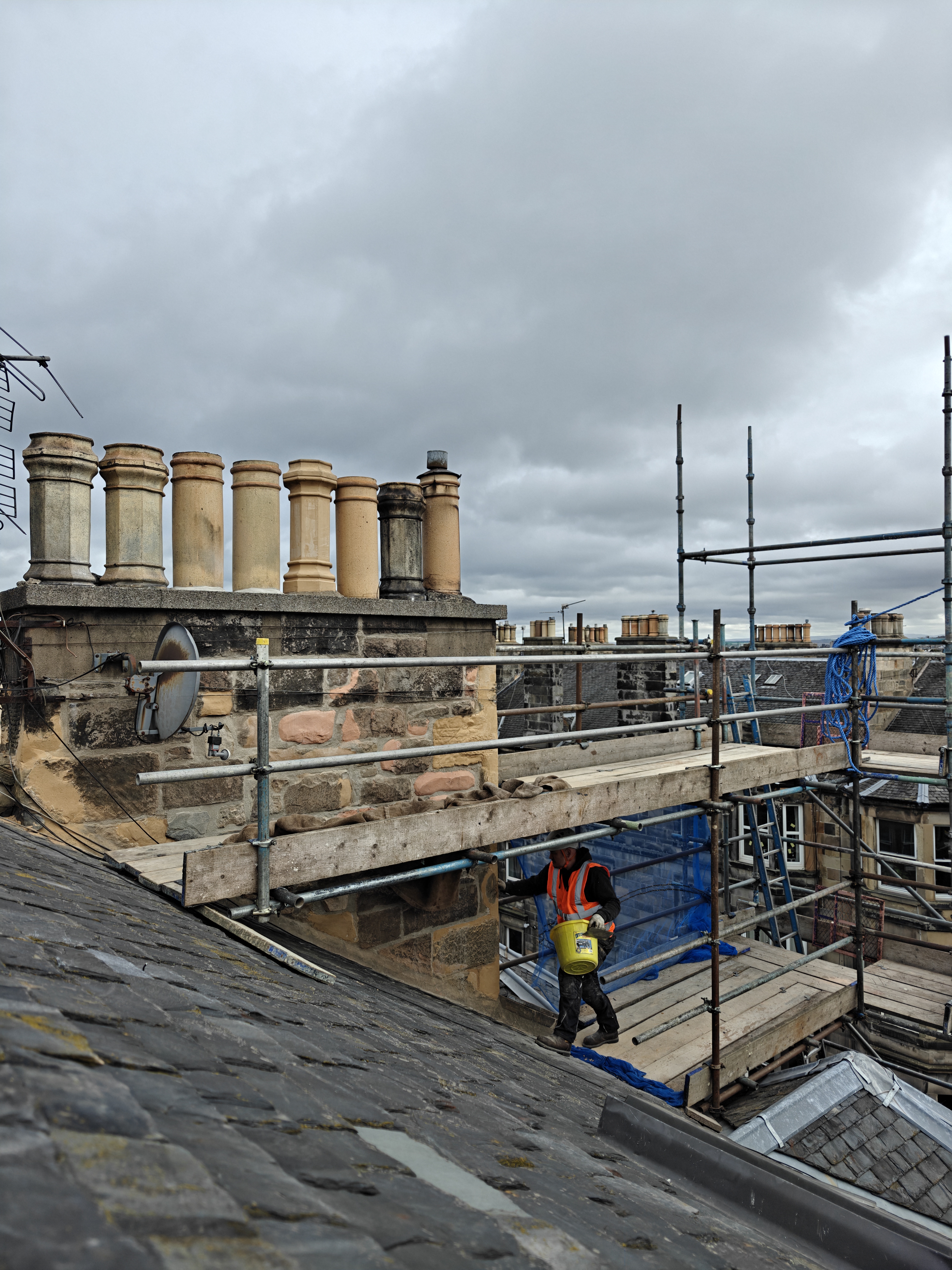 A worker in a high-visibility vest walks on a sloped roof, carrying a yellow bucket, with chimney stacks and scaffolding visible in the background under a cloudy sky.