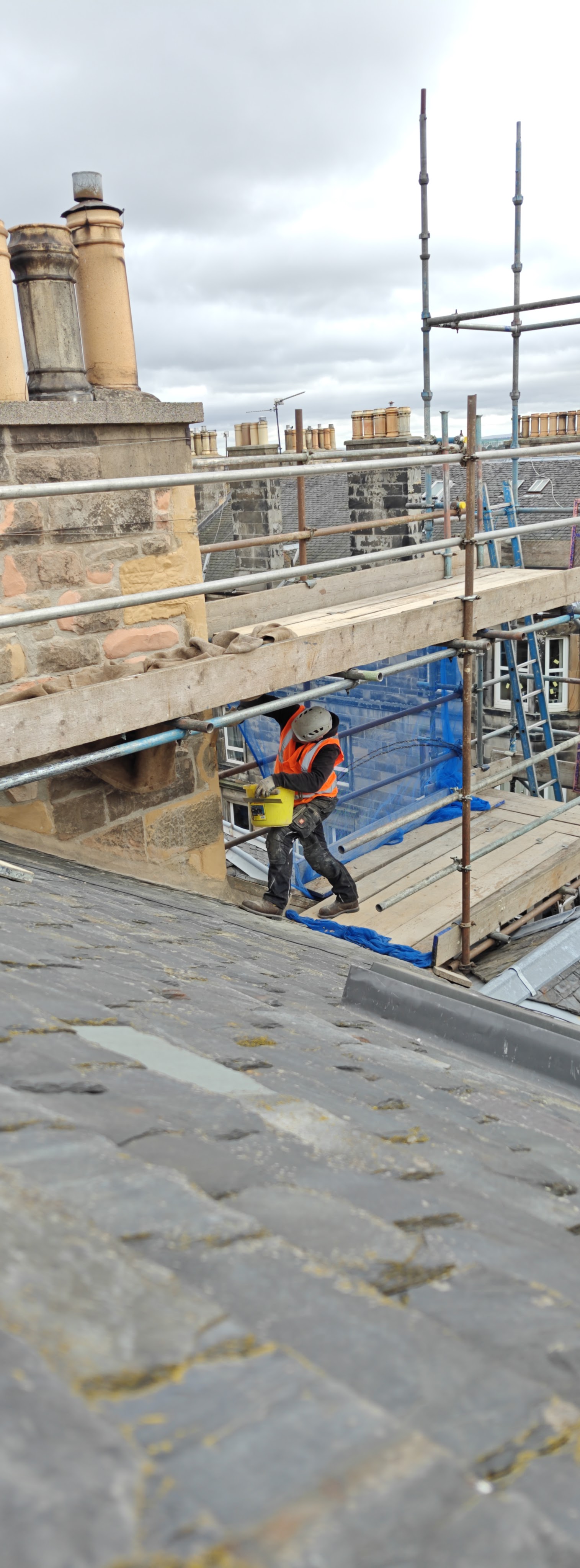 A construction worker in a safety vest and helmet carries a yellow bucket while navigating a sloped roof, with scaffolding and chimney stacks visible in the background.