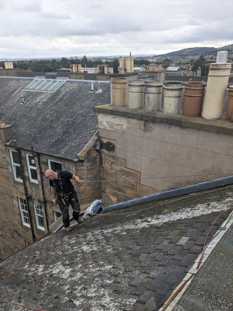A worker dressed in safety gear is inspecting a sloping roof covered with slate tiles, surrounded by chimneys on a cloudy day, with a view of nearby buildings and hills in the background.