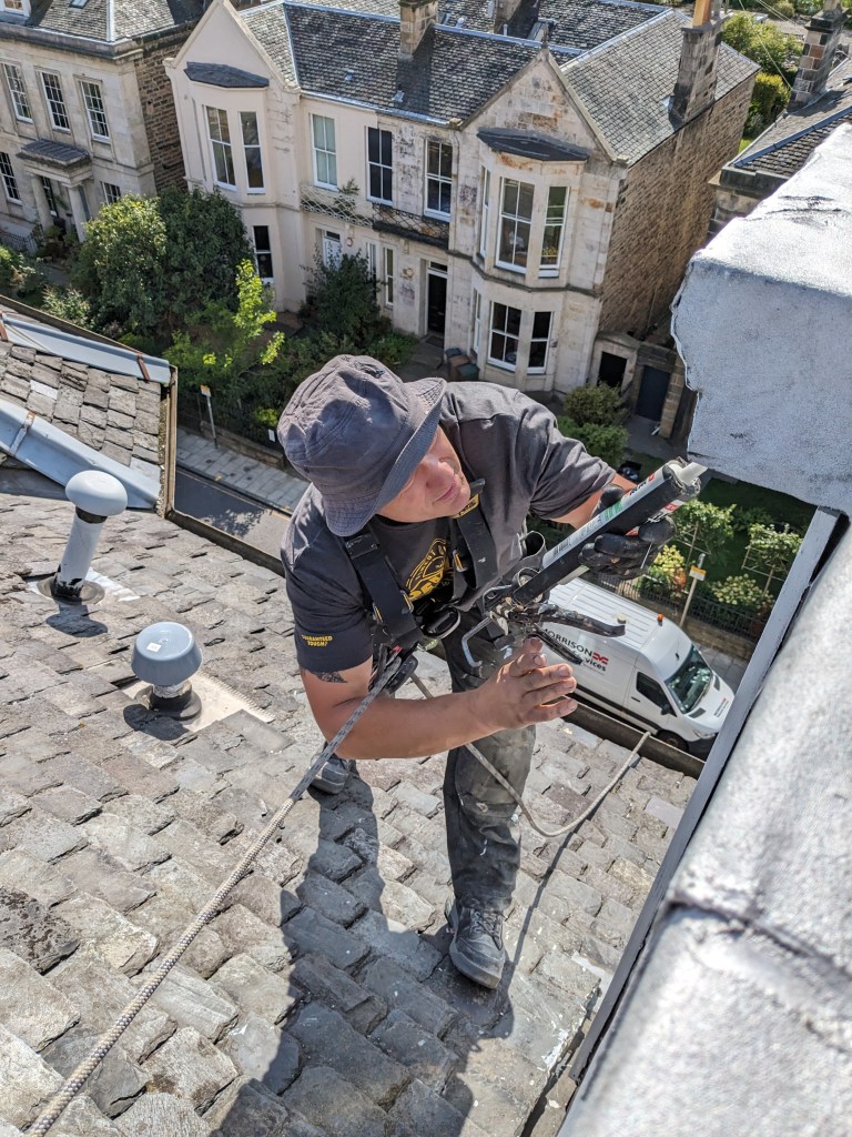A worker using safety harnesses and equipment to perform maintenance on a sloped roof, with residential buildings visible in the background.