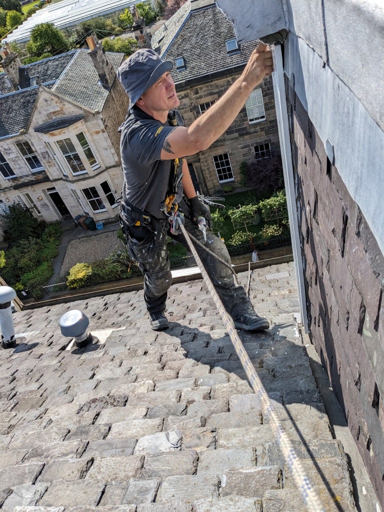 A roofer working on a steep slate roof, using safety harnesses and ropes, with residential buildings and greenery visible in the background.