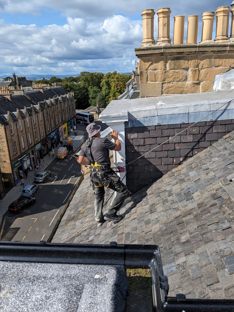 A roofer working on a sloped roof, adjusting equipment while overlooking a street lined with buildings and trees under a cloudy sky.
