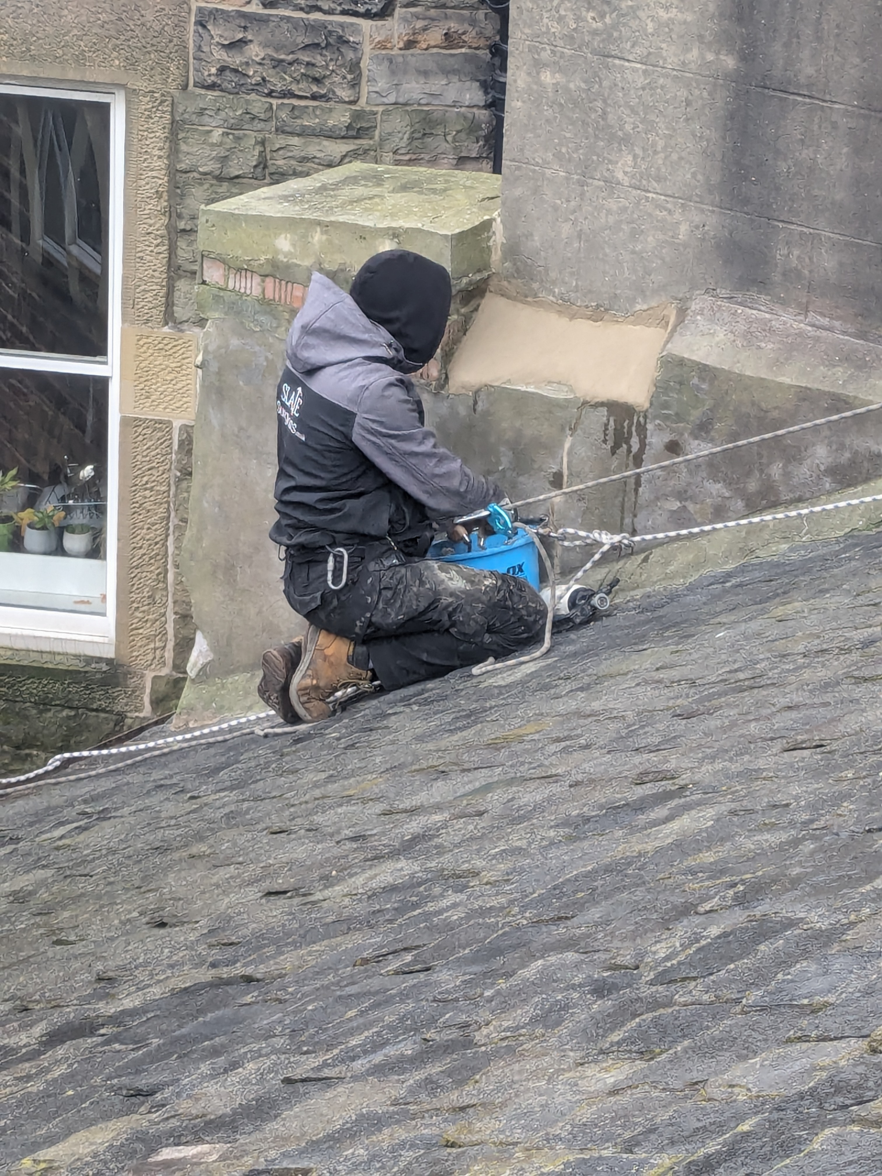 A roofer wearing a black hoodie and safety gear is using a power tool on a sloped roof, with a stone building in the background.