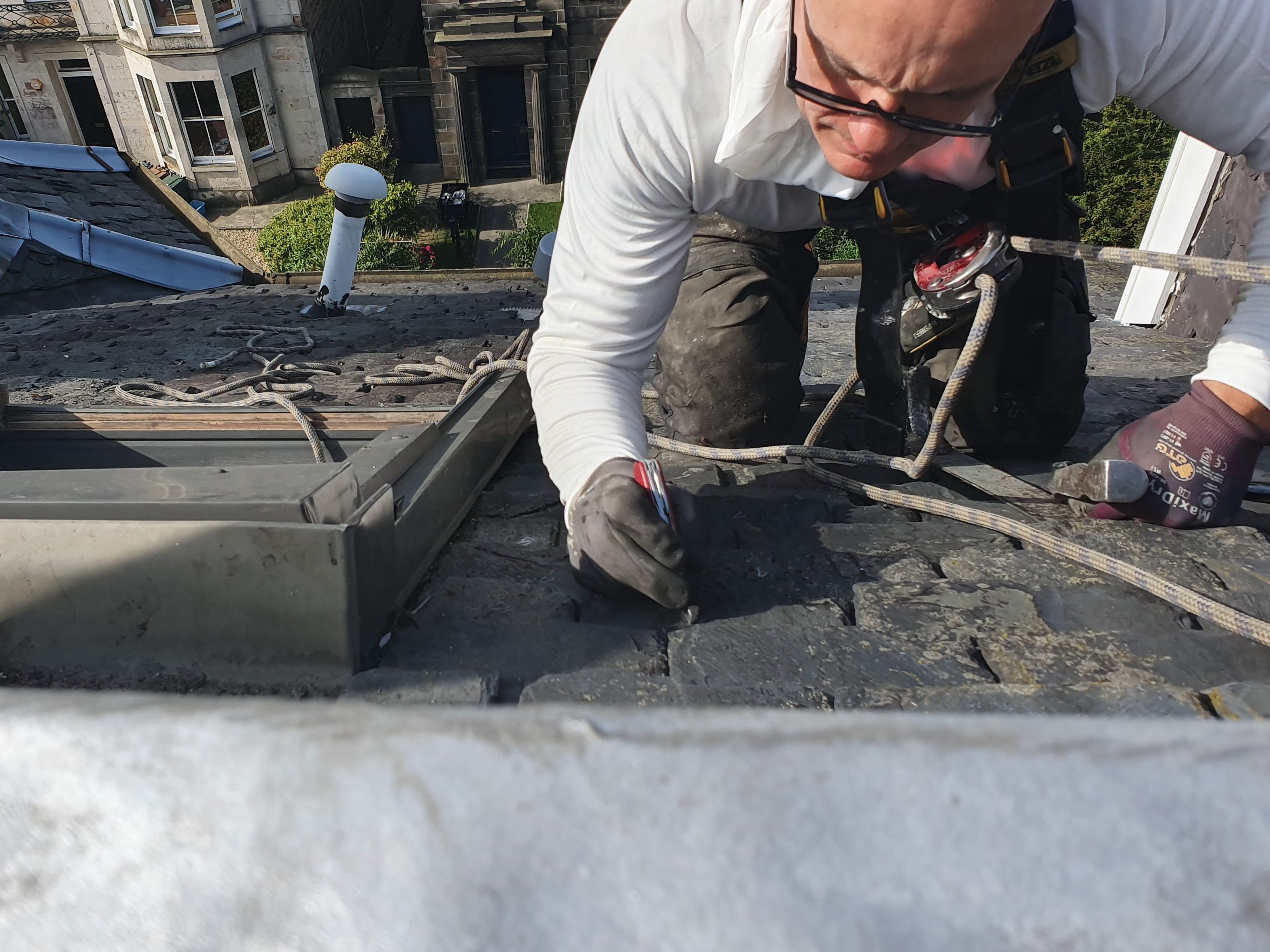 A person wearing safety gear is working on a roof, using tools to make repairs on slate tiles while secured with safety ropes.